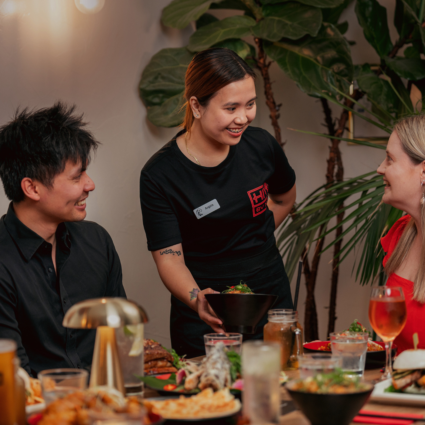 Three people pictured, focus is on the staff member serving Asian inspired dishes to a blonde female and dark hair male.